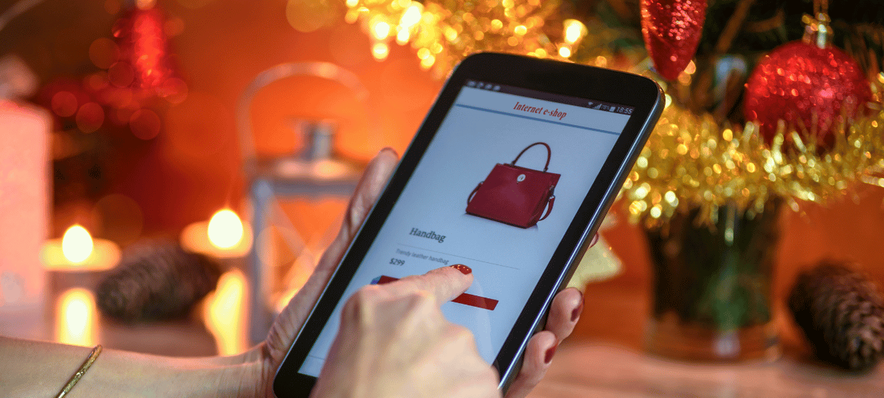 Na obrázku je v pozadí vánoční stromeček a vládne vánoční atmosféra. V popředí obrázku je osobota držící tablet a nakupuju online na tabletu.