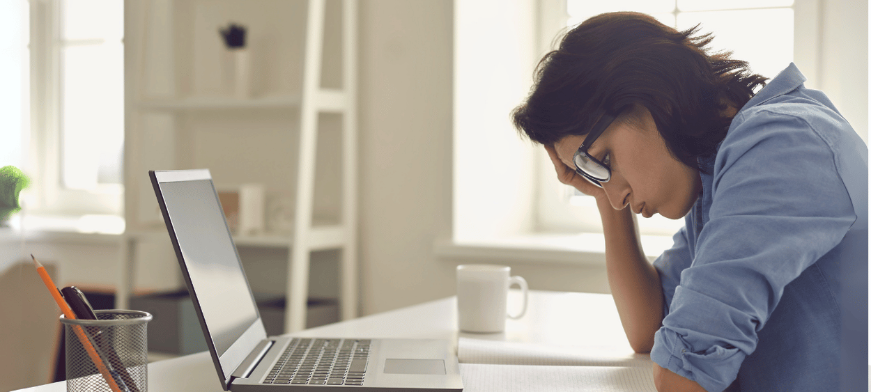 Pohled z boku na ženu sedící u počítače. Drží se za hlavu a vypadá unaveně a zoufale.