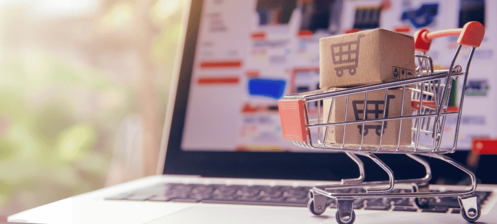 Atrapa/ hračka nákupního košíku položeného na klávestini notebooku. V nákupníkm košíku jsou dané mále krabice