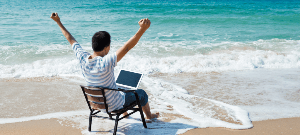 muž s počítaček na klíně sedící na židli na pláži a moře u šplouchá u nohou.